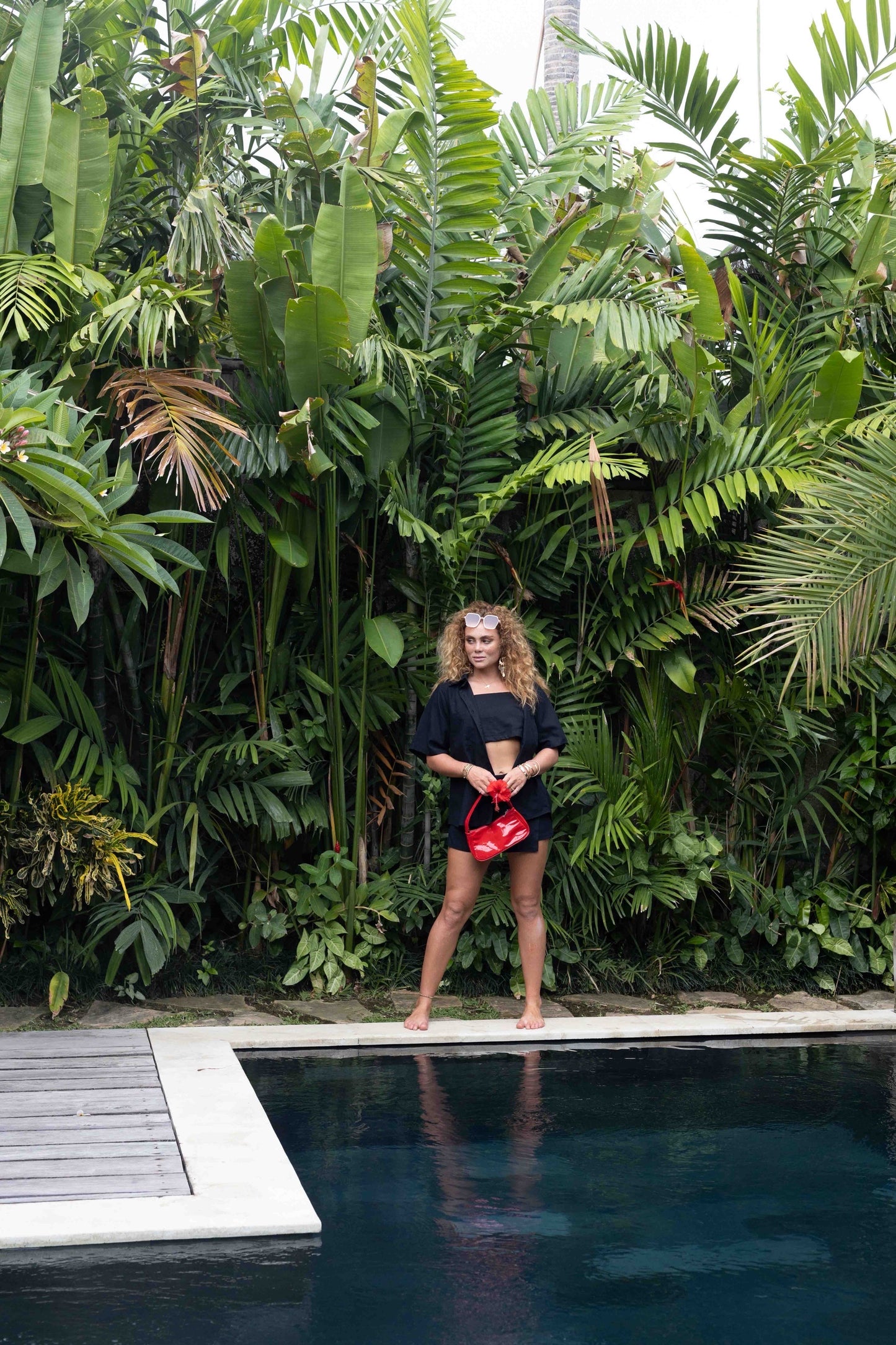 Woman wearing black beach shirt for women by MOSS The Label standing near pool with tropical plants