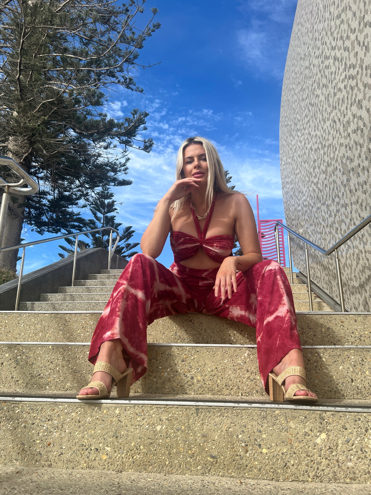 Woman wearing red tie-dye jumpsuit by MOSS The Label sitting on outdoor stairs against blue sky