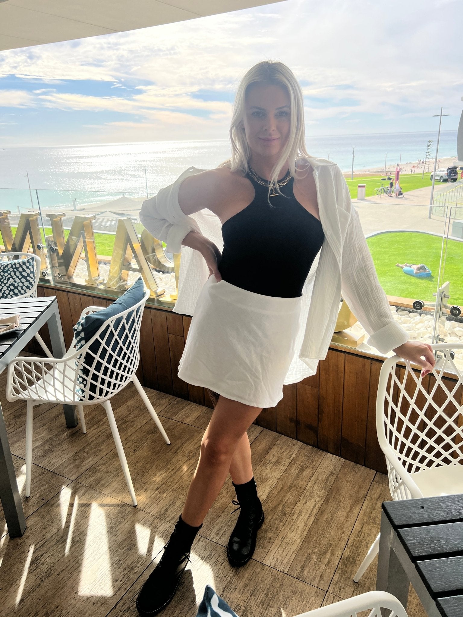 Woman wearing a black bodysuit with a white skirt and jacket standing by a seaside cafe